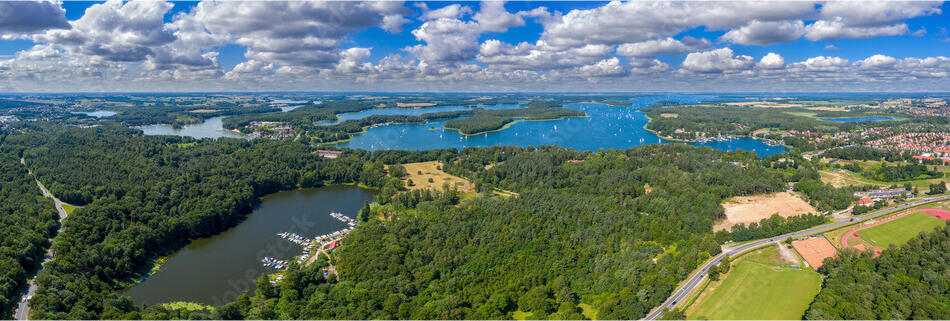 Landscape photograph. Aerial view of lakes, forests, and distant buildings, with the sky partially covered with clouds above.