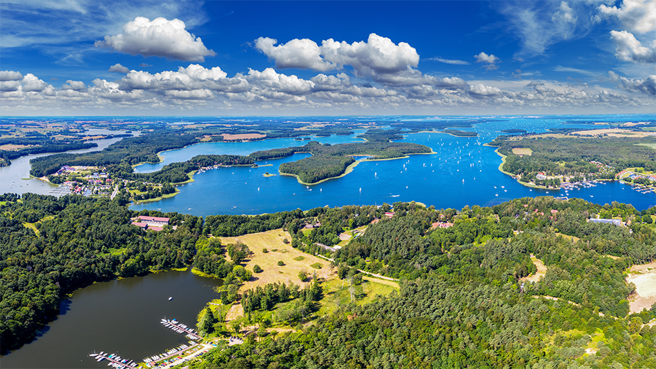 Landscape photograph. Aerial view of lakes, forests, and distant buildings, with the sky partially covered with clouds above.